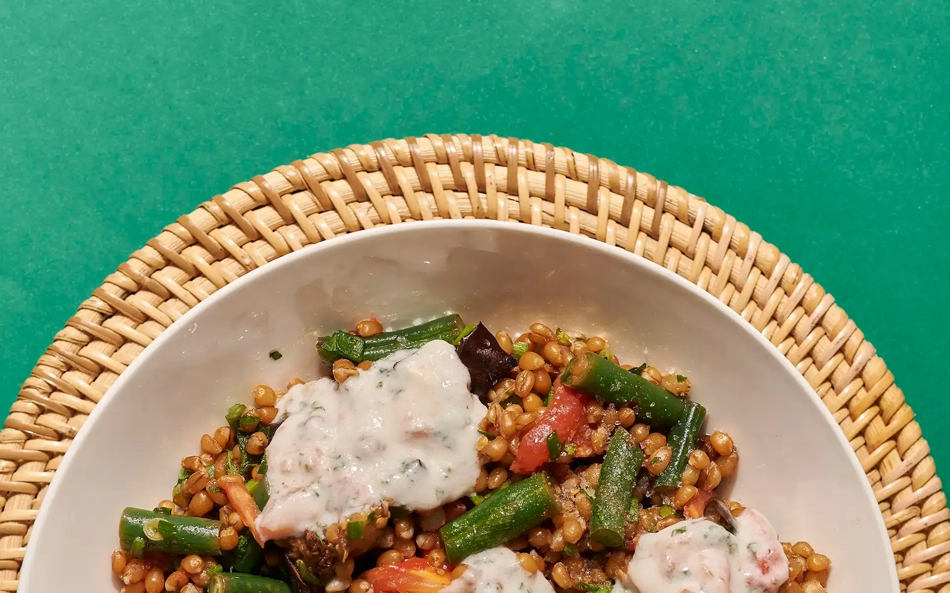Salada de grãos (tipo trigo) com vagem, tomate e molho de iogurte, em tigela de porcelana sobre suporte de palha, fundo verde